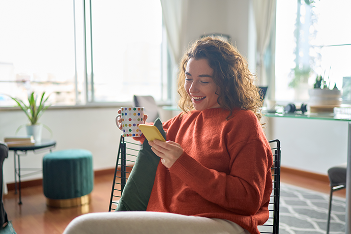 Smiling woman holding smartphone drinking tea relaxing at home